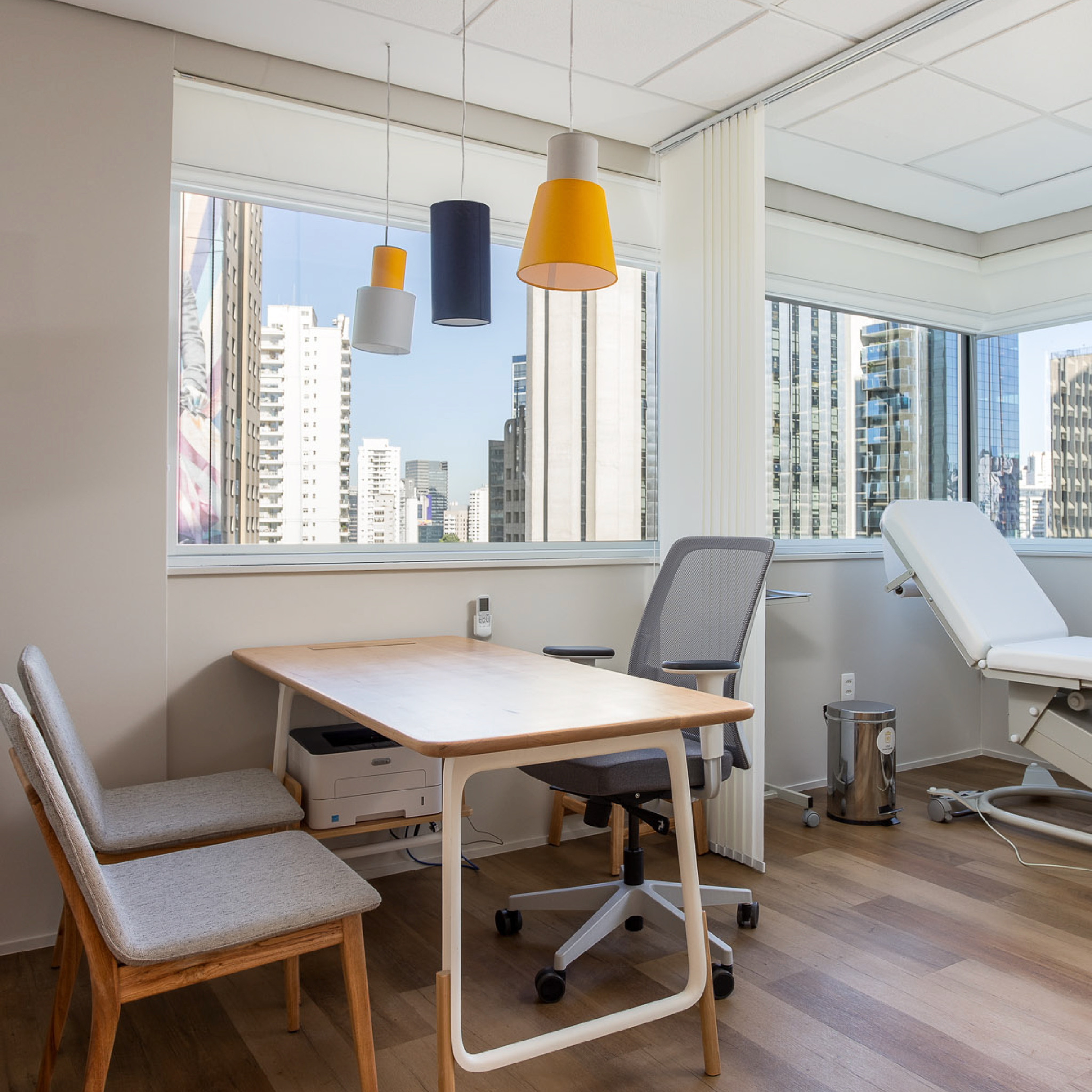 Consultório com mesa de atendimento, cadeiras e maca, iluminado por janelas